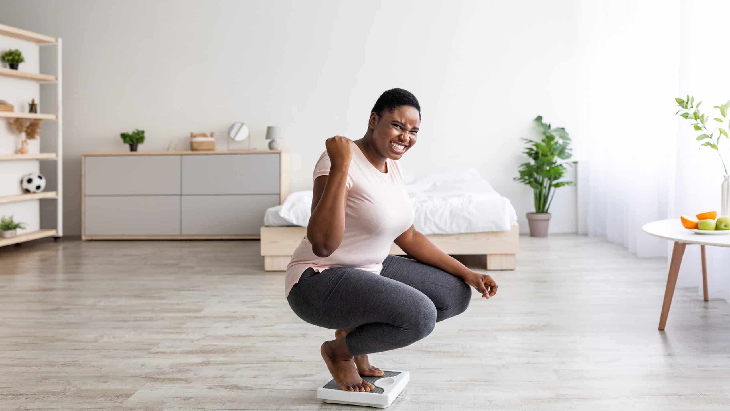 Excited black woman sitting on scales at home, making YES gesture, happy with result of her slimming diet